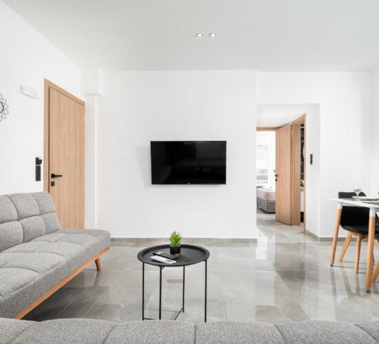 Bright modern living room with a grey sofa, small round coffee table, wall-mounted TV, and a view into the dining area and bedroom at ELEANA Beach Suites.