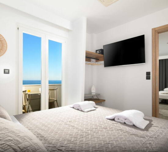 Modern bedroom featuring a double bed with fresh linens, a balcony overlooking the sea, a wall-mounted TV, and an open wardrobe at ELEANA Beach Suites