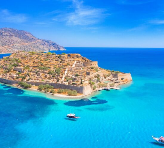 Aerial view of Spinalonga Island in Crete with turquoise waters, historic fortifications, and boats surrounding the shore.