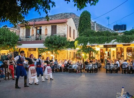 Traditional Cretan dance night in a village square with dancers in traditional costumes and guests dining at a local taverna.