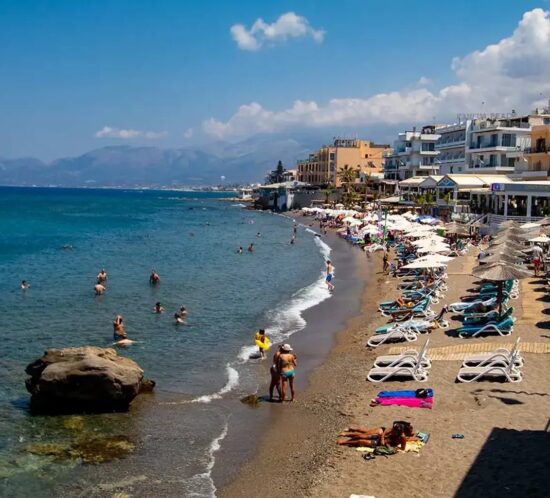 Strand in Chersonissos auf Kreta mit Sonnenliegen, kristallklarem Wasser und Besuchern, die ihr Bad unter der sommerlichen Sonne genießen.